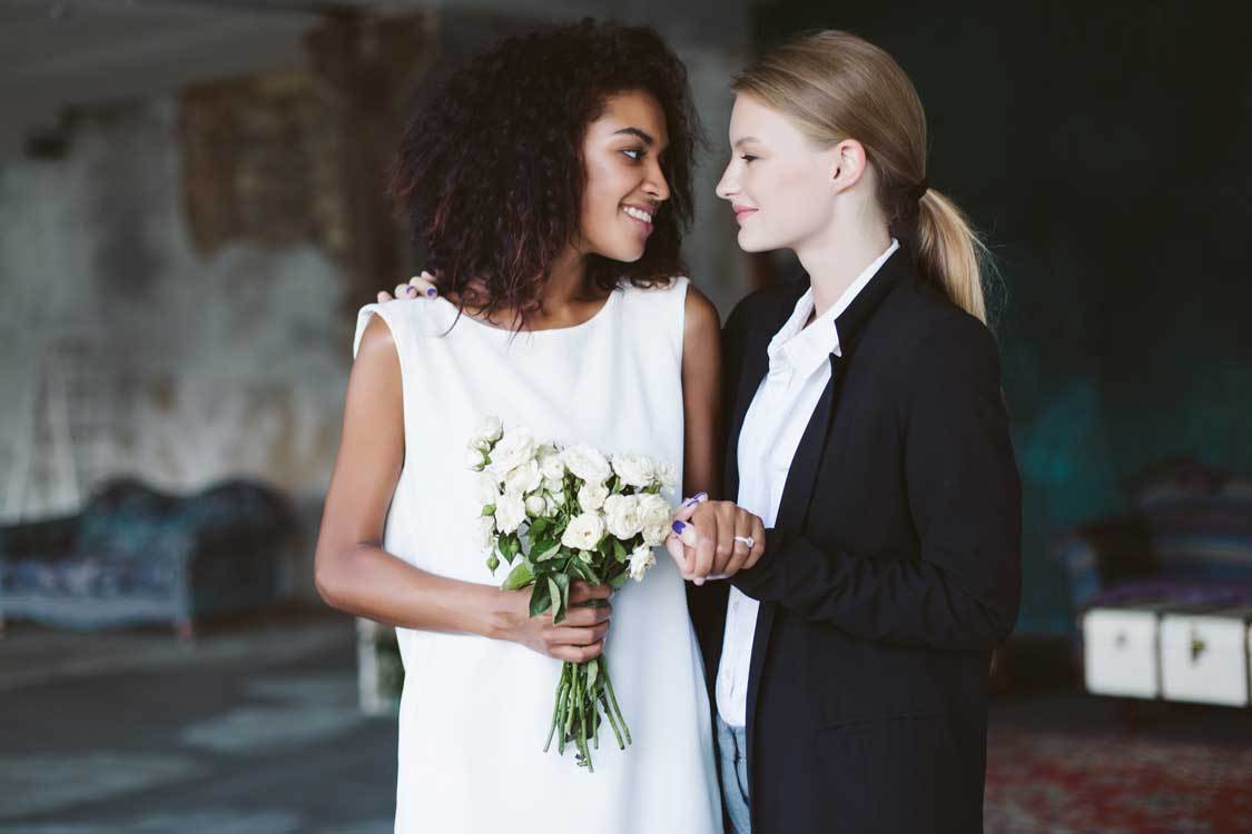 Young woman with blond hair in black suit and smiling african american woman with dark curly hair in white dress with bouquet of flowers in hand happily looking at each other on wedding